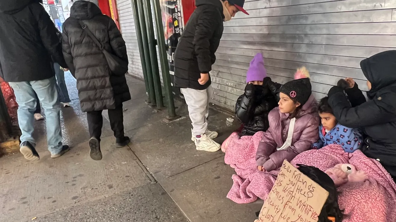 Children and adults wrapped in pink blankets sit on a city sidewalk against a closed metal shutter; one holds a handwritten sign asking for help while passersby walk by.