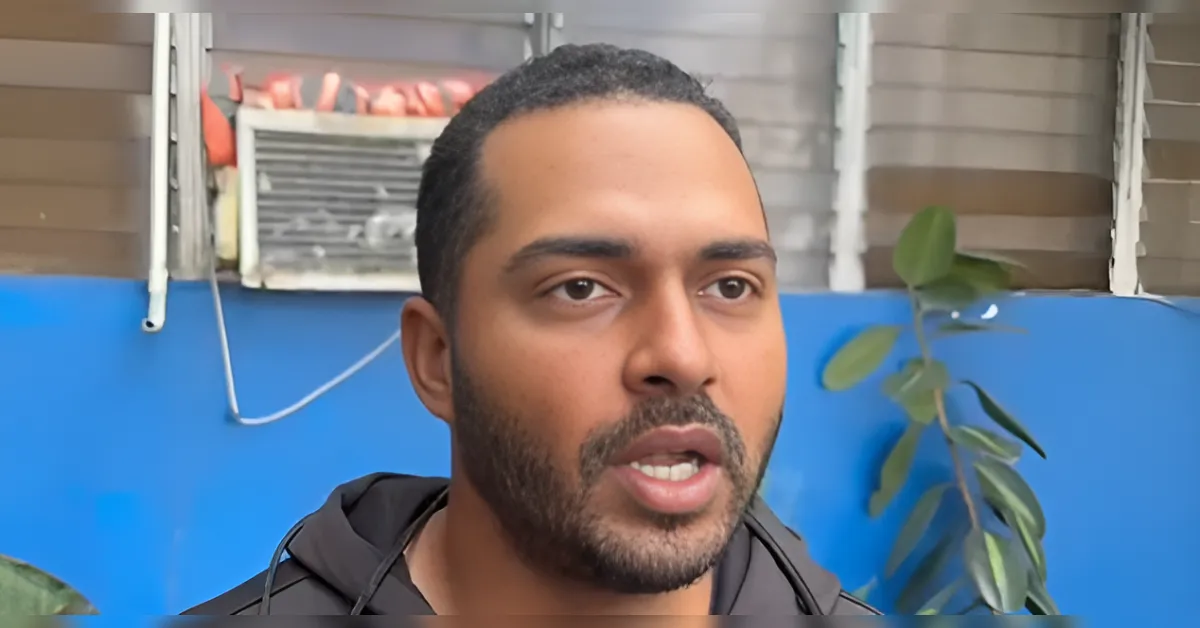 Man with short hair and beard speaks, indoor setting with blue wall and plants in the background.