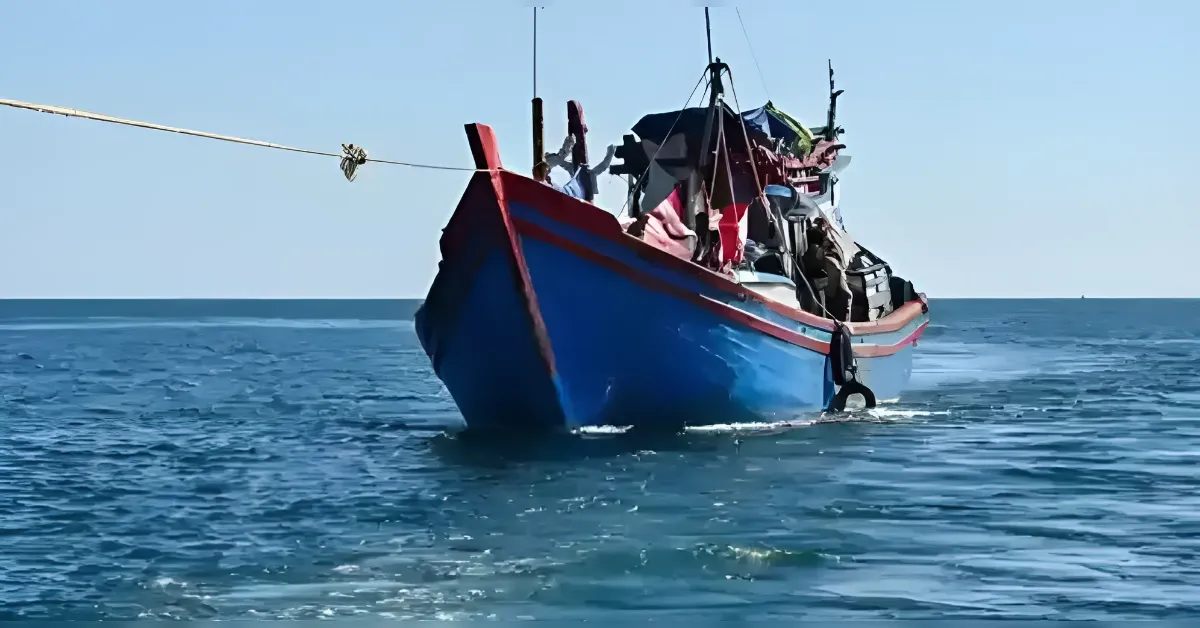 Blue fishing boat with red trim floating on calm sea under a clear blue sky, with people on the deck visible.