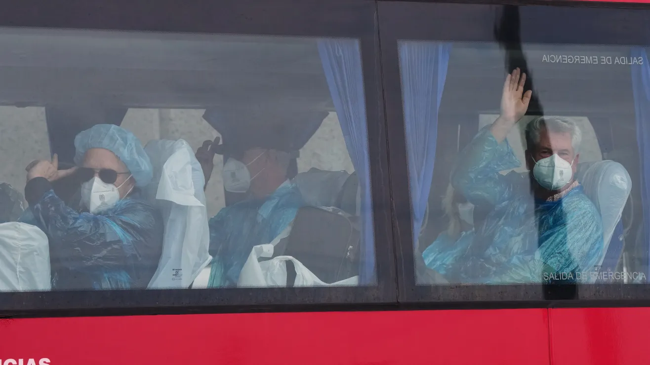 People in protective medical gear and masks inside a red bus, waving from the windows; emergency exit text visible on the glass.