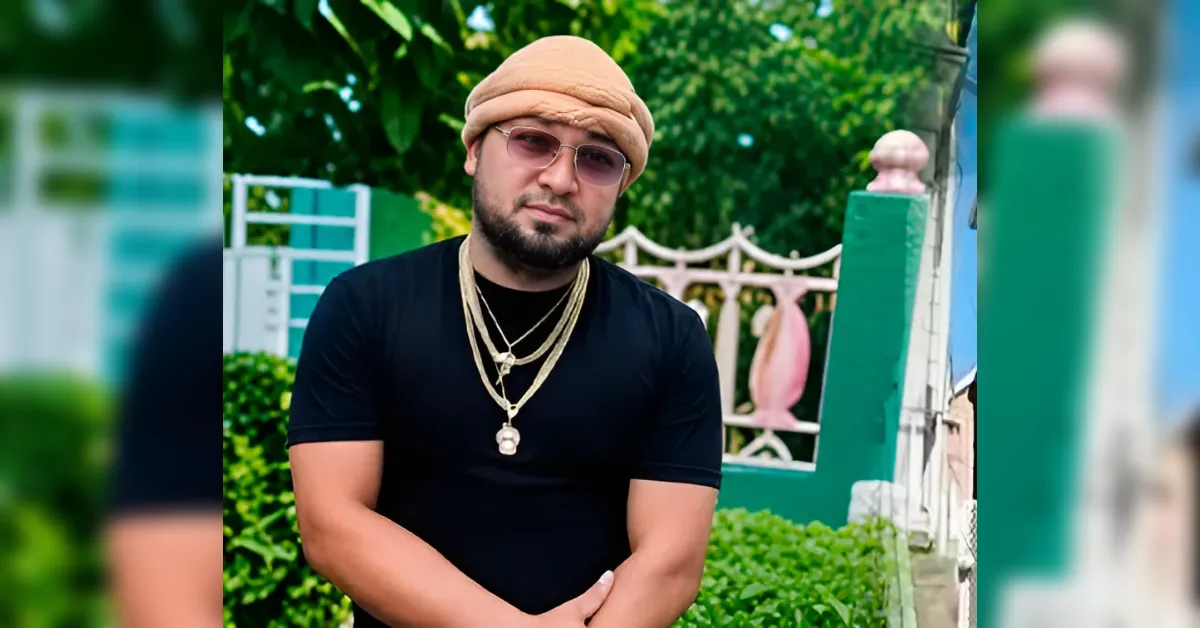 Man wearing a tan turban, pink-tinted glasses, and layered gold chains posing outdoors near a green fence and hedge.