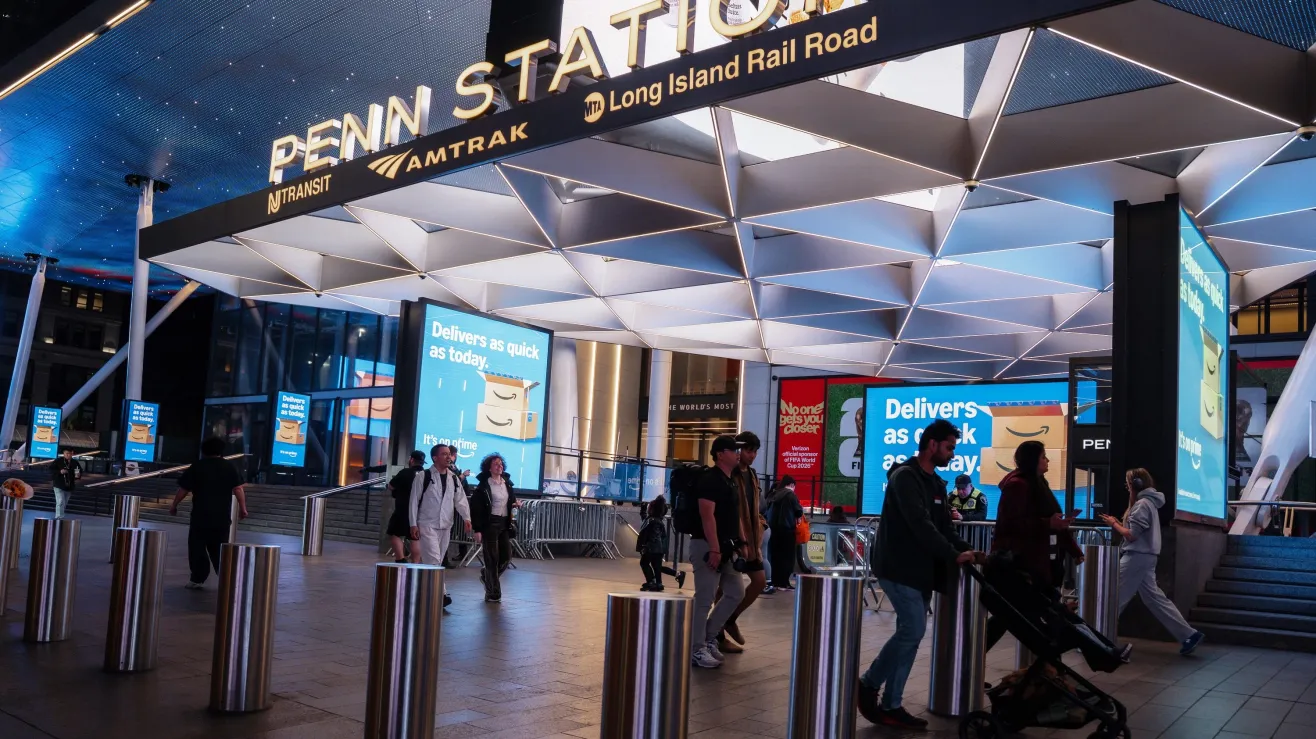 Busy Penn Station concourse with a geometric white ceiling, large Amtrak and Long Island Rail Road signs, and travelers walking past metal bollards and digital billboards.