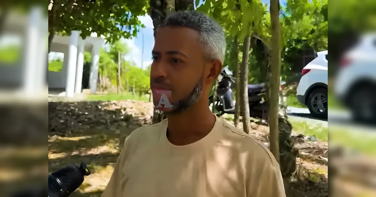Man being interviewed outdoors, speaking to a microphone in a sunny, tree-lined area.