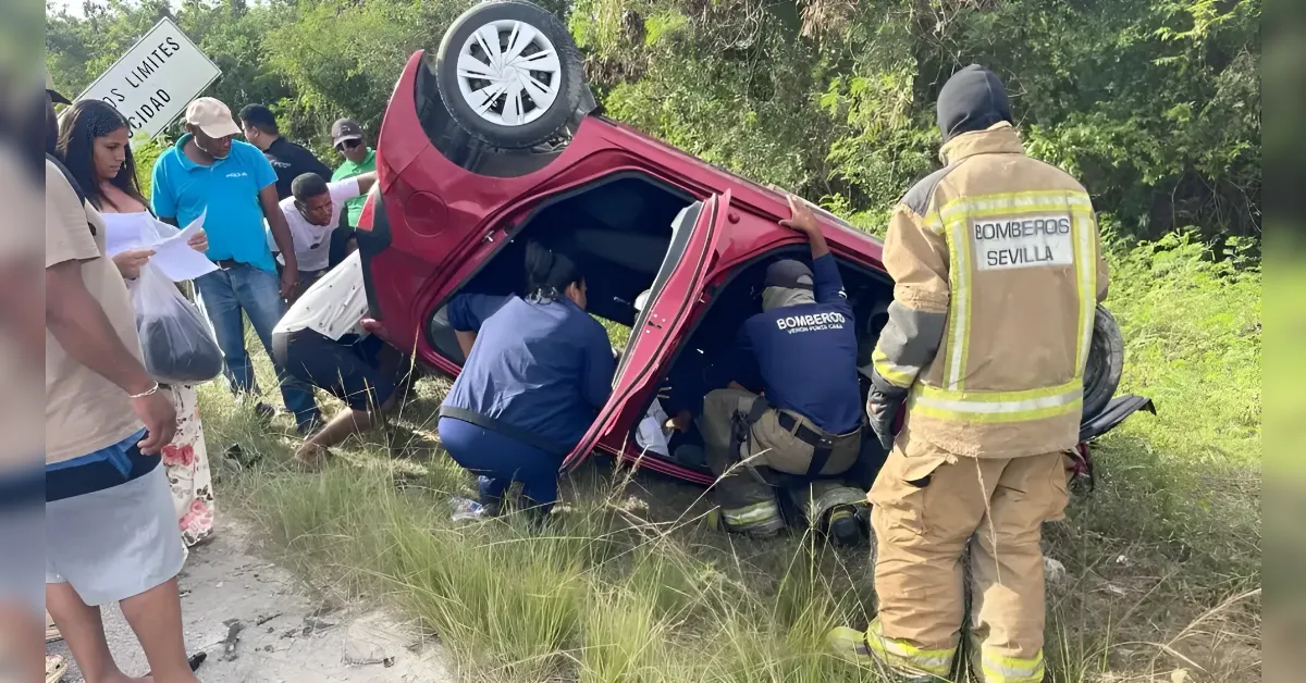 Emergency responders in blue uniforms and a firefighter in beige gear extricate occupants from a red car flipped onto its side in a grassy roadside area.