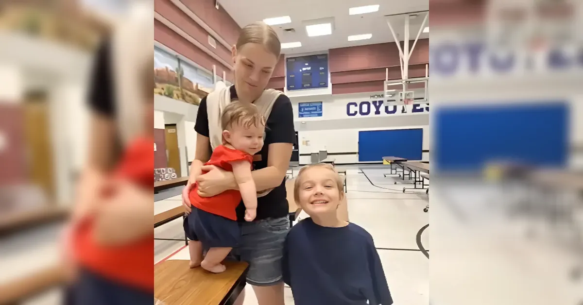 Mother holding a baby in red near her arm, with an older child smiling beside them in a school gymnasium