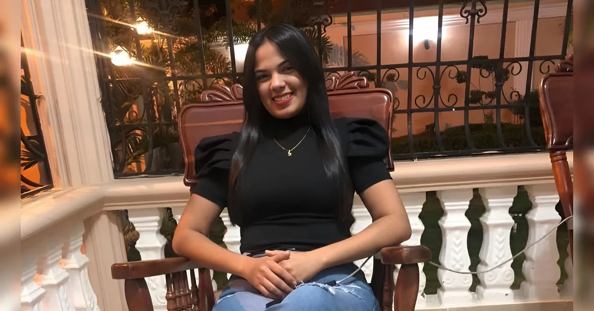 Woman with long dark hair sits on a wooden chair on a veranda, smiling at the camera, wearing a black top and blue jeans.