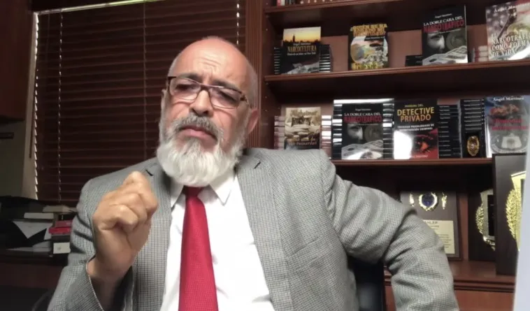Older man with gray beard and glasses in a gray suit and red tie, seated in an office with bookshelves behind him, gesturing as he speaks.