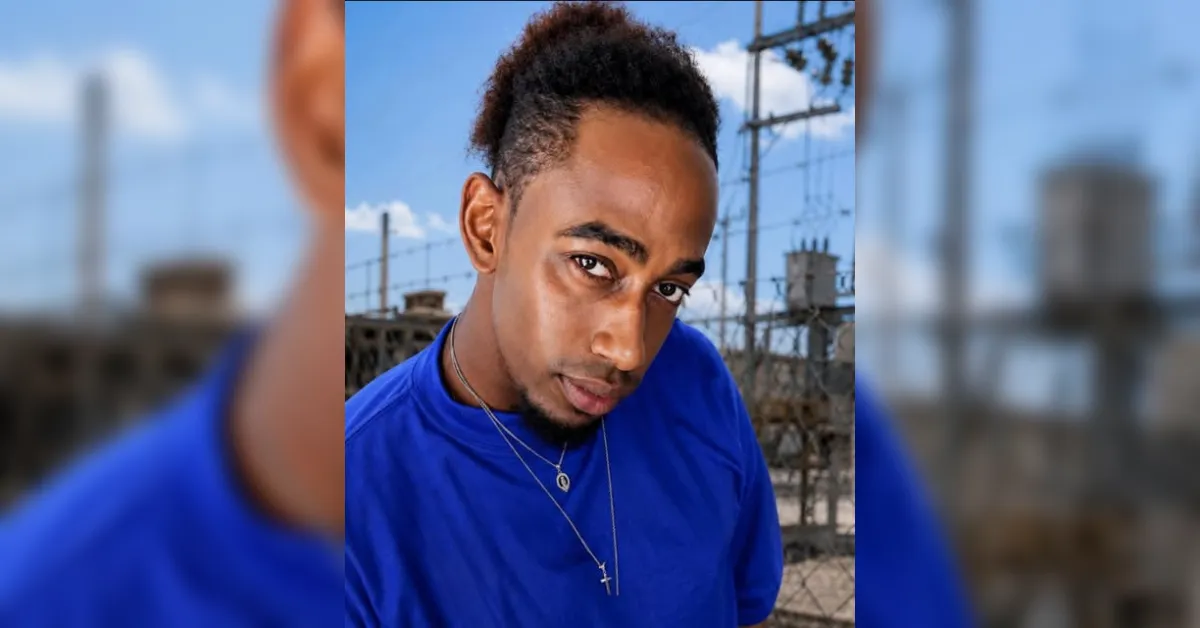 Close-up portrait of a young man in a bright blue shirt, wearing two necklaces, looking into the camera with a serious expression, outdoor setting with power lines in the background
