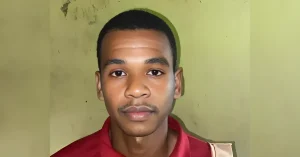 Portrait of a young man with short black hair wearing a red shirt, standing against a pale green wall.