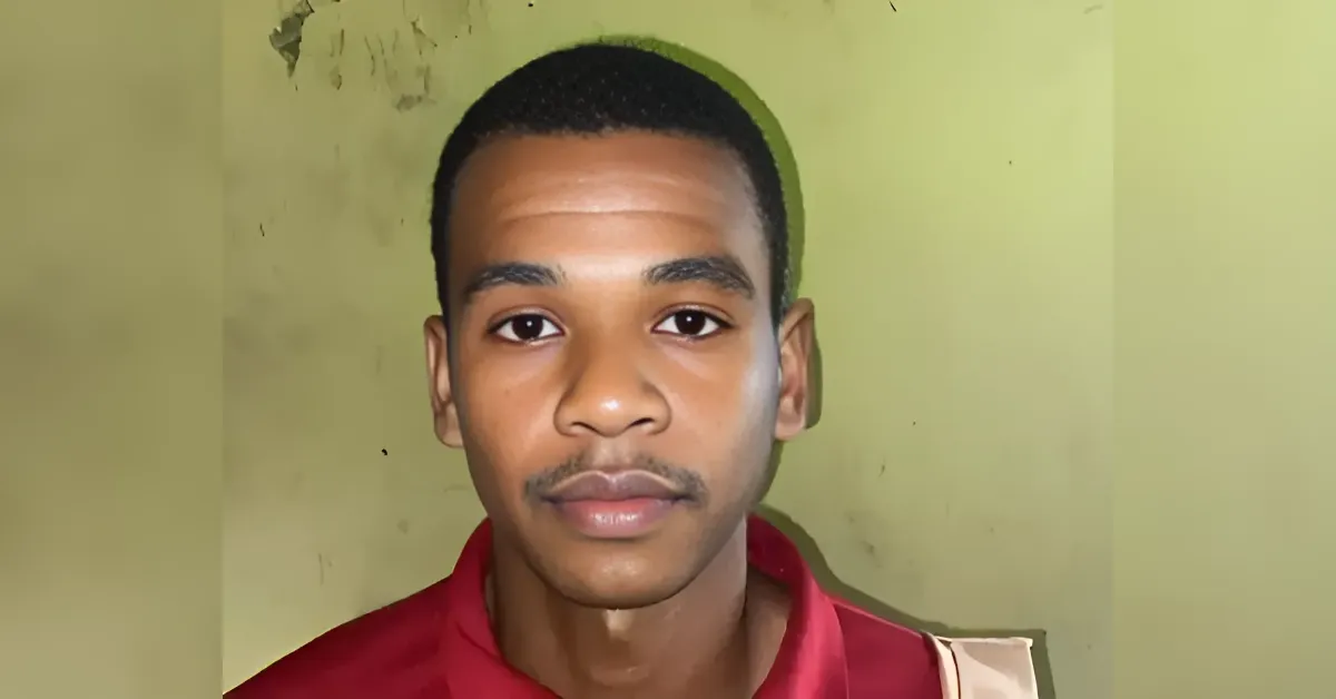 Portrait of a young man with short black hair wearing a red shirt, standing against a pale green wall.