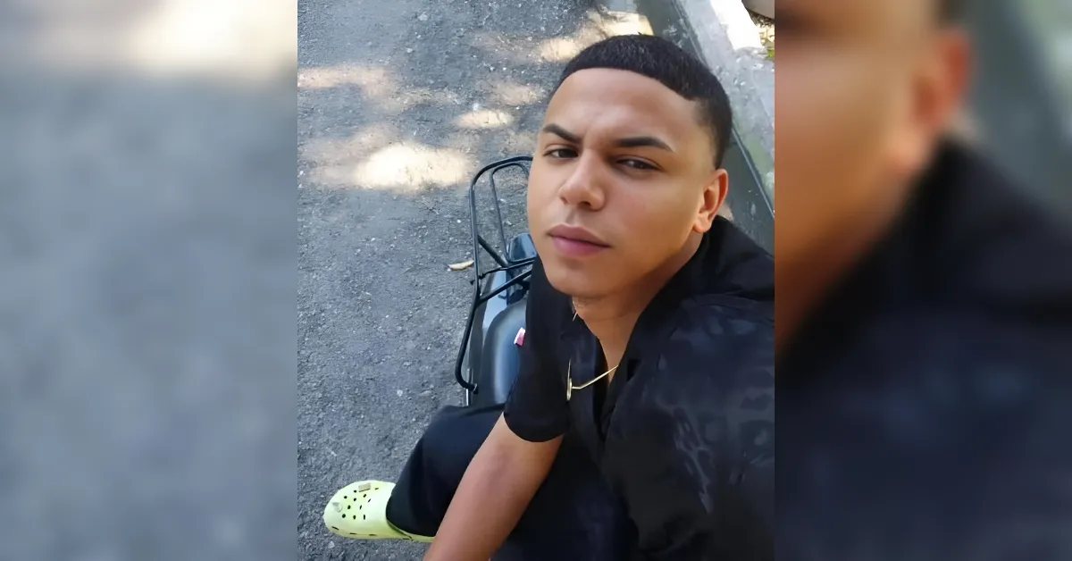 Young man in a black shirt with a gold chain sits on a scooter outdoors, looking at the camera.