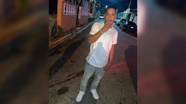 Young man in a white t-shirt, light jeans, and white sneakers stands on a dimly lit street at night, his hand near his mouth.