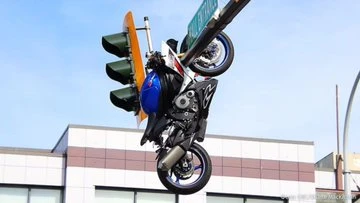 Motorcycle stunt rider performs a hang-trick on a traffic light above a city street, wearing a helmet and protective gear.
