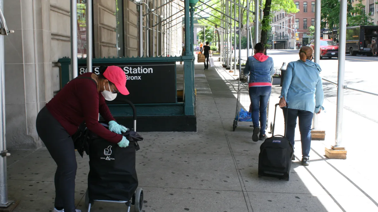Woman wearing a pink cap and mask pushes a wheeled cart with a black cover along a city sidewalk near a station entrance labeled 'Bronx.' A second person pulls a small backpack on wheels in the background as a street runs with cars and pedestrians.