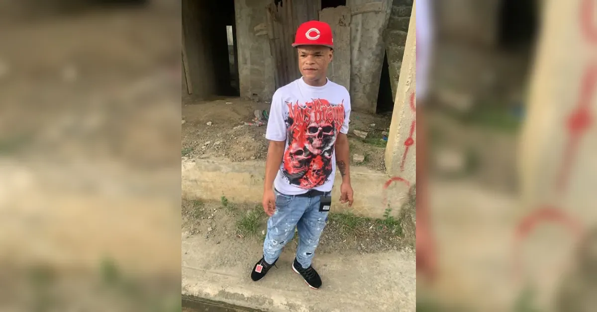 Young man standing on a dirt path in front of an unfinished building, wearing a white graphic t-shirt, light blue ripped jeans, black sneakers, and a red cap.