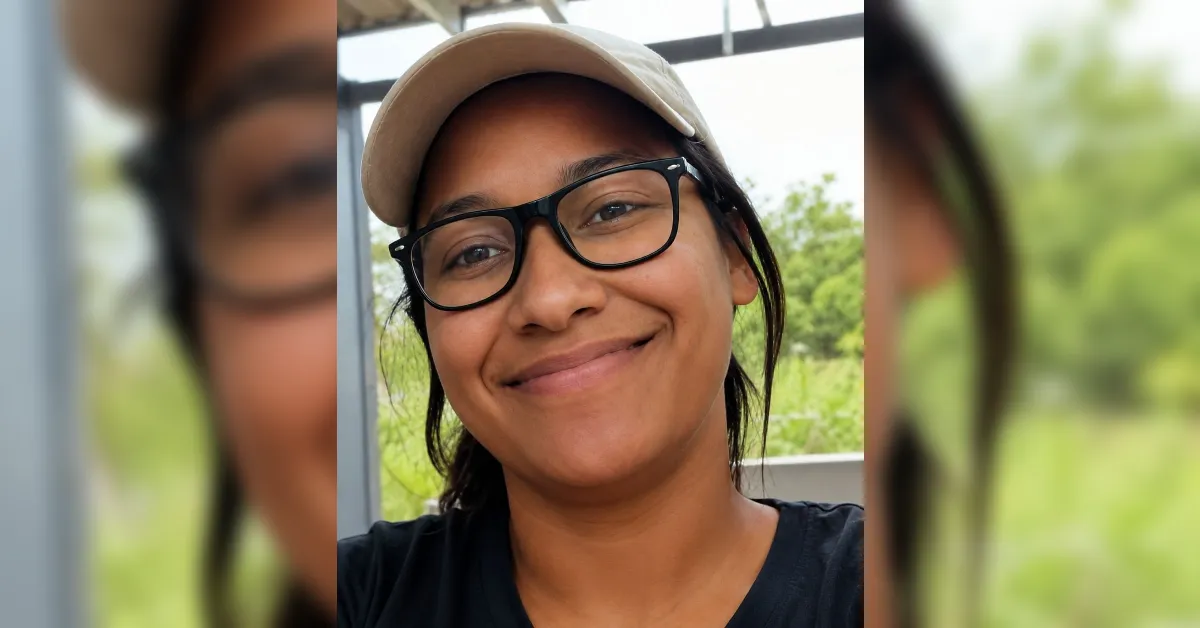 Smiling woman outdoors wearing black-framed glasses and a beige cap (selfie).
