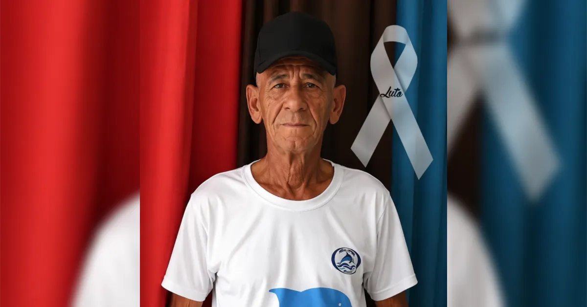 Older man in a white T-shirt and black cap stands before red and blue drapes with a white awareness ribbon.