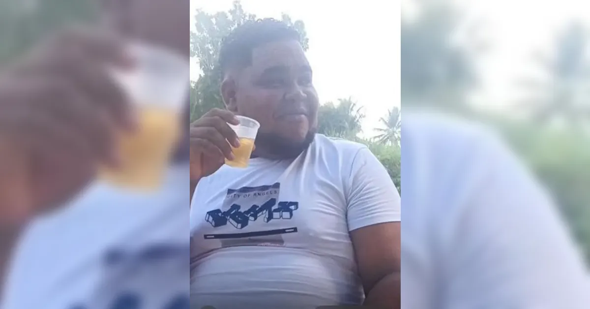 Man smiling and raising a plastic cup of beer outdoors, wearing a light gray t‑shirt.