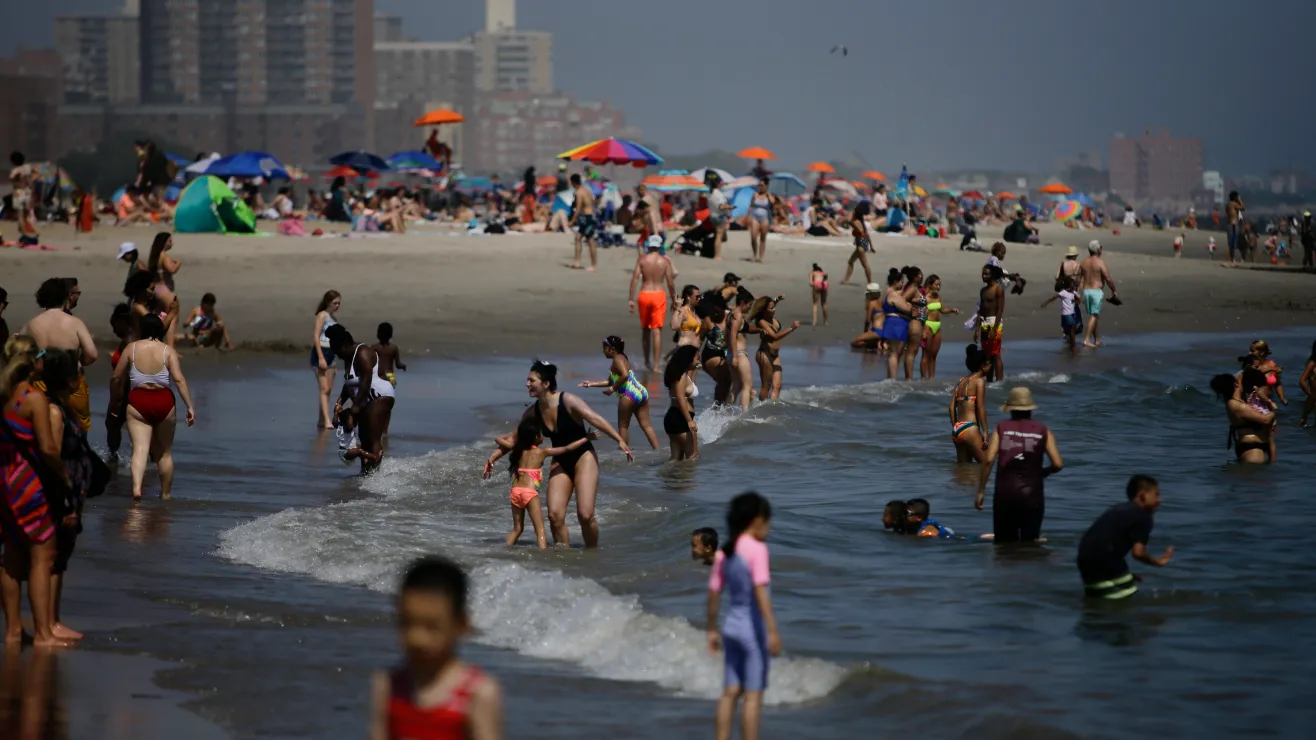 Crowded beach with families and swimmers in shallow water; colorful umbrellas and buildings in the background.