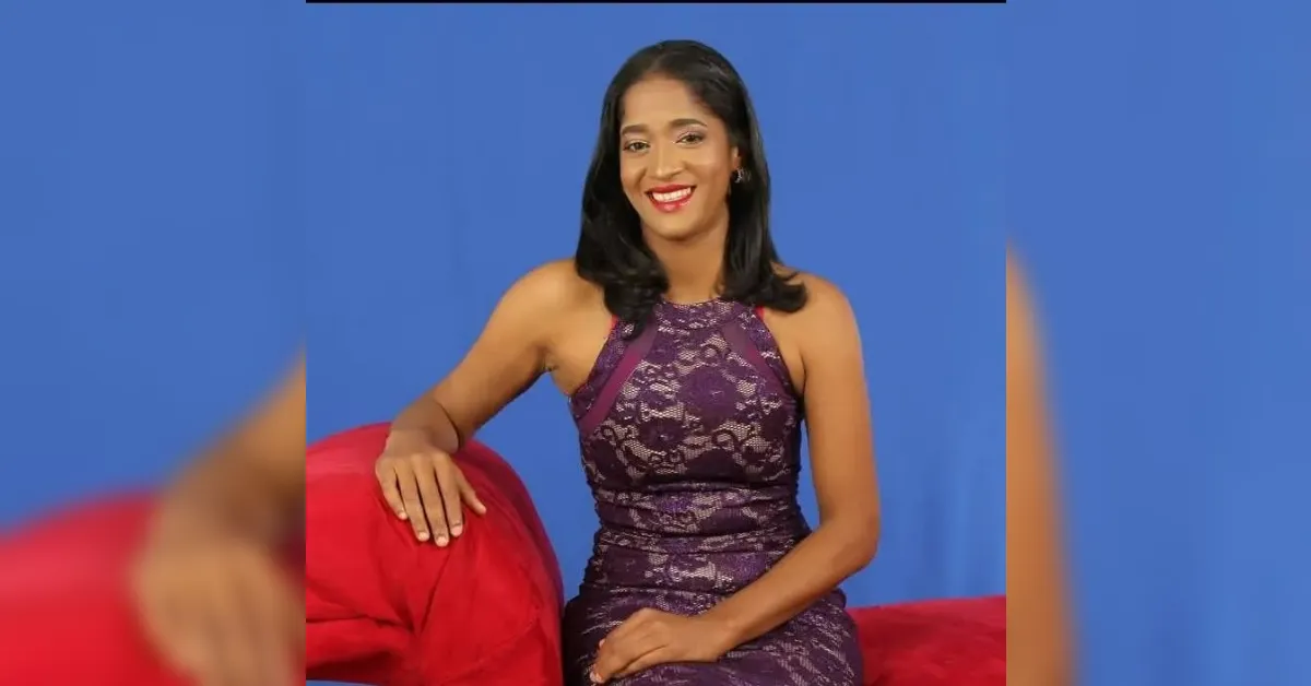 Woman in a purple lace evening dress sits on a red couch against a blue backdrop, smiling at the camera.