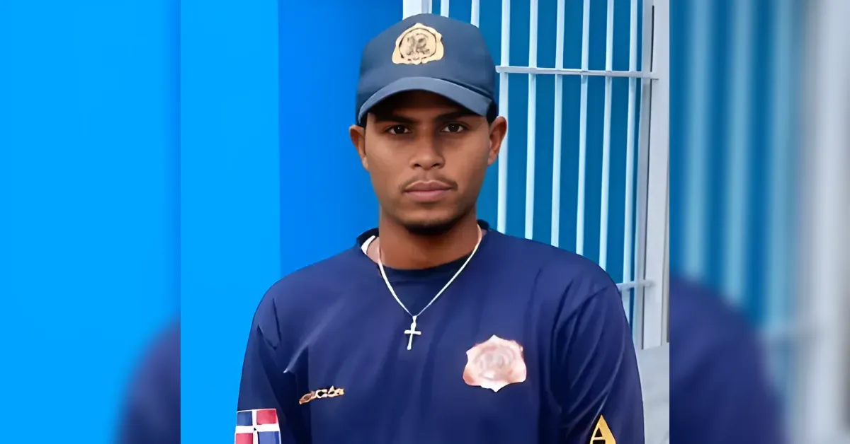 Young man wearing a dark blue shirt with a rose patch and a cap, standing in front of a blue wall with vertical bars, wearing a cross necklace.