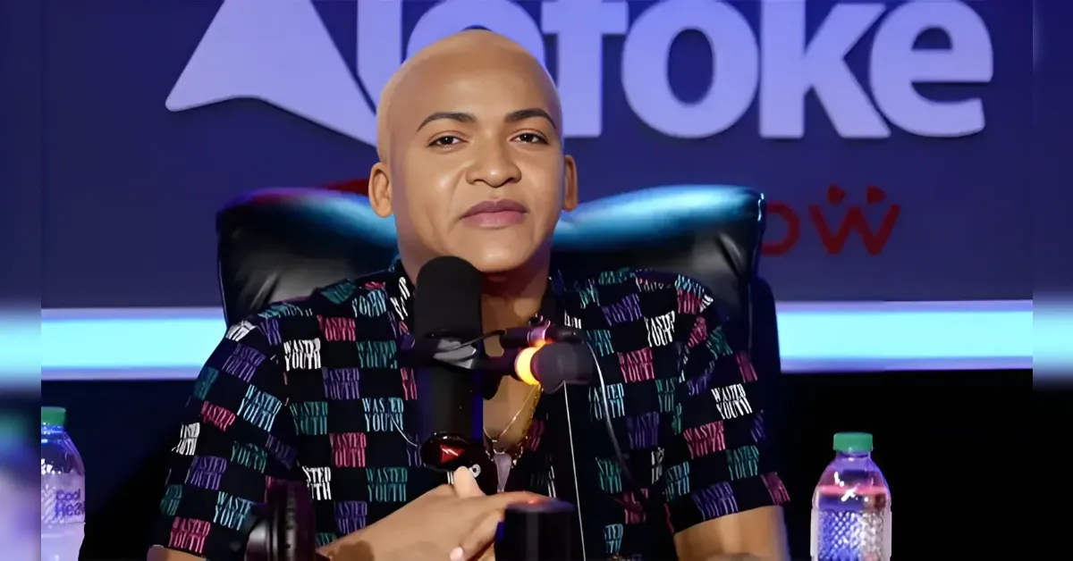 Person with a shaved head speaking into a microphone at a panel, wearing a colorful patterned shirt with water bottles on the desk in front of a blue backdrop.