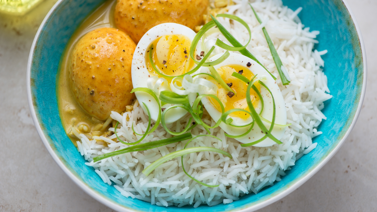 Blue bowl of white rice topped with halved hard-boiled eggs, scallions, and yellow curry with potatoes.