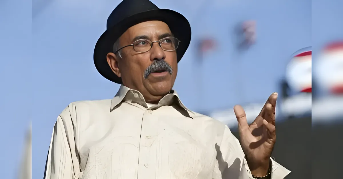 Older man with a mustache and glasses wearing a black fedora speaks outdoors, gesturing with his right hand against a blue sky background.