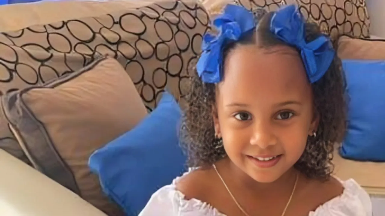 Young girl with curly hair wearing blue bows and a white off-shoulder top, smiling on a living room couch.
