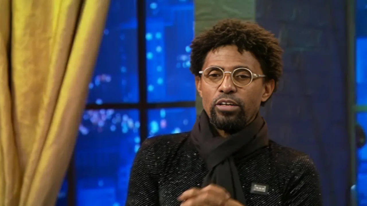 Man with round glasses and a dark scarf sits in a TV studio with blue city lights in the background.