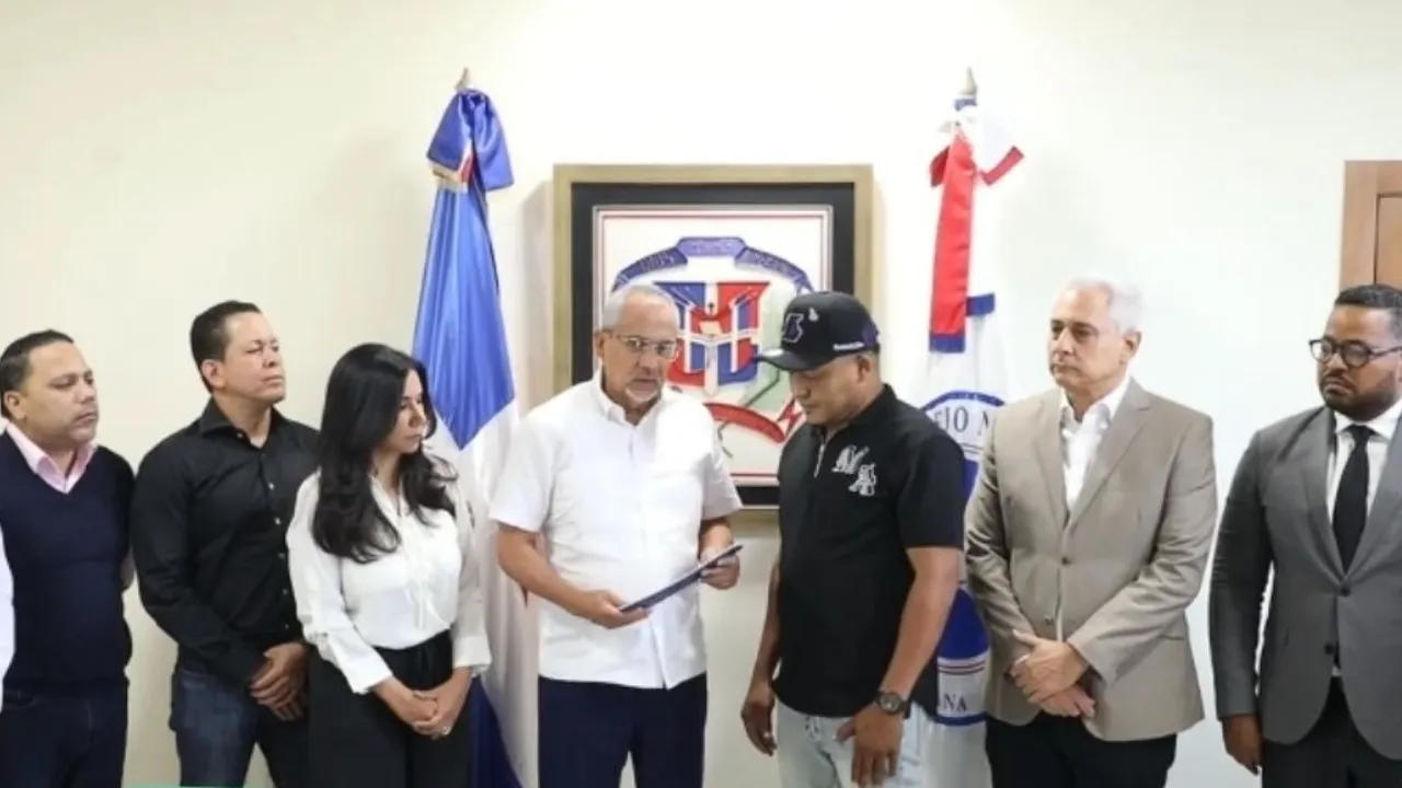 Man in a white shirt hands a folder to a man in a black polo while several colleagues watch in a room with flags and an emblem on the wall