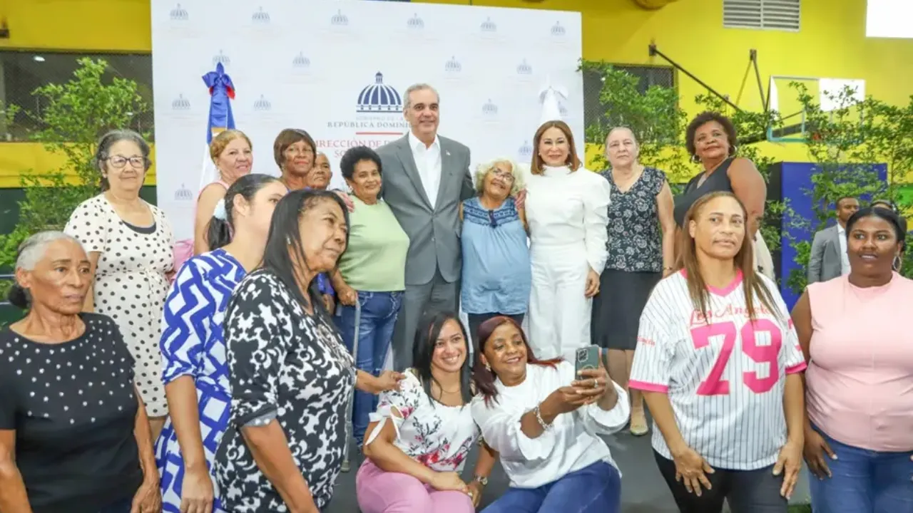 Group of diverse women and a man in a gray suit posing for a photo at a formal event, with a Dominican Republic emblem backdrop behind them.