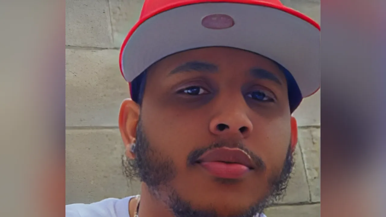 Close-up selfie of a young man wearing a red and gray cap, with a trimmed beard, looking at the camera.