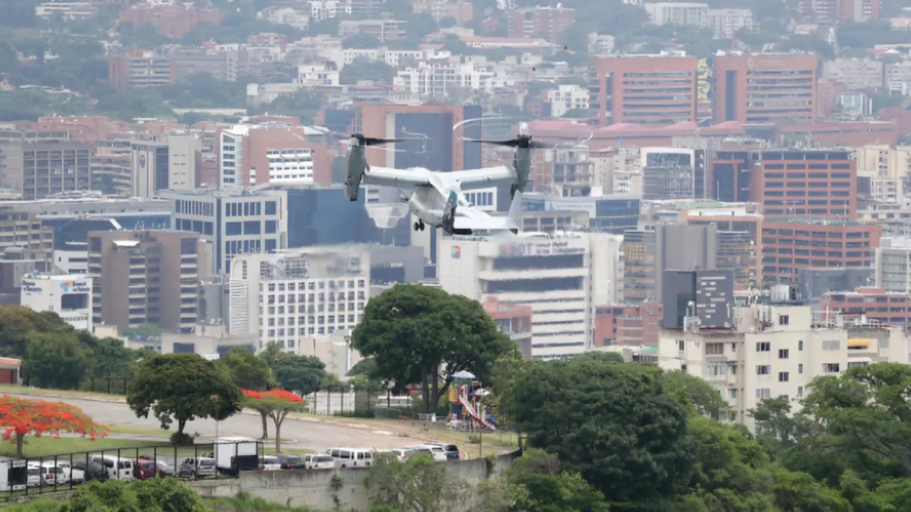 White quadcopter drone hovering over a cityscape with tall buildings and green trees in the foreground