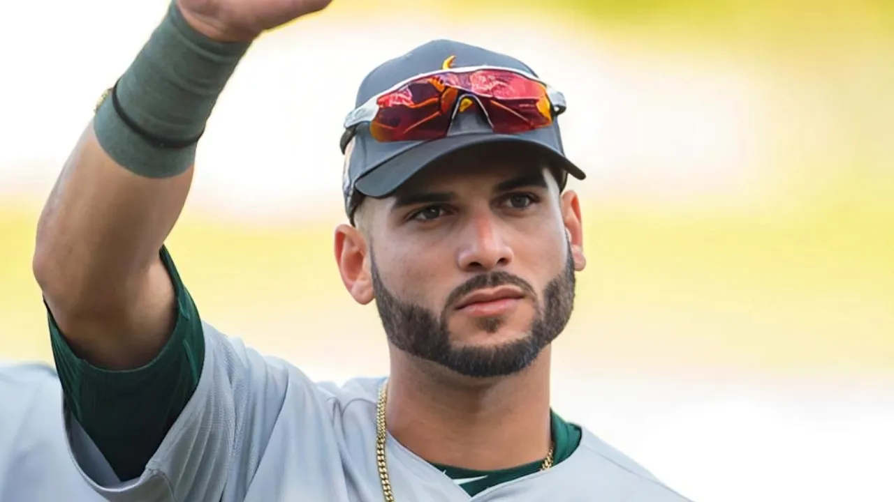 Athletic man with beard wearing a gray baseball jersey and cap, raising his hand outdoors with red sunglasses on his cap.