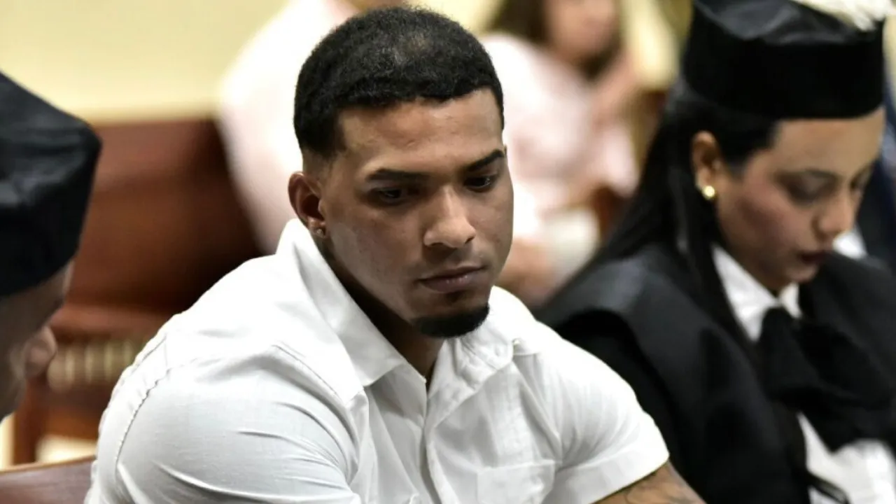 Man in a white shirt seated in a courtroom, looking downward, with a graduate in cap and gown in the background.