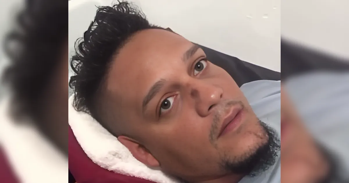 Close-up of a man lying on a bed with a white pillow behind his head, looking at the camera