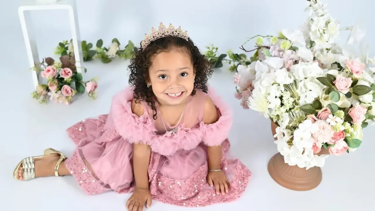Young girl in a pink princess dress and tiara kneels on a light blue studio floor beside a large bouquet of white and pink flowers.