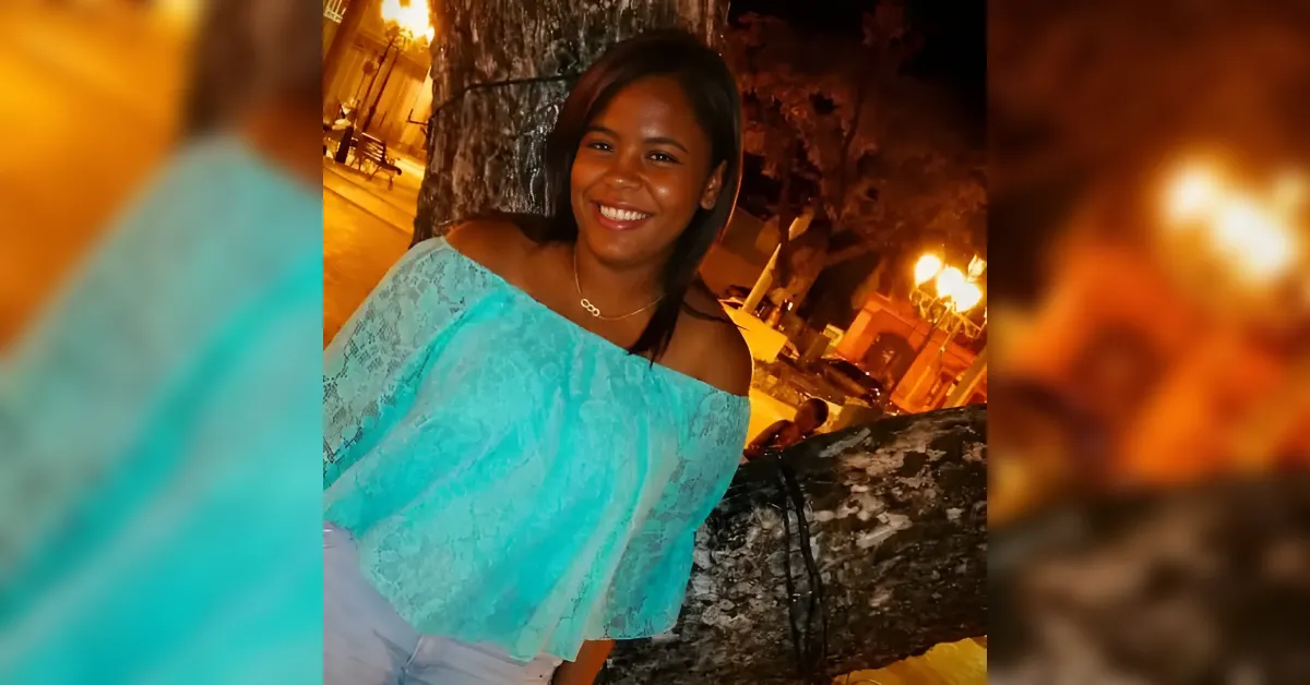 Smiling woman wearing a turquoise off-the-shoulder top poses beside a tree at night with warm torchlight in the background.