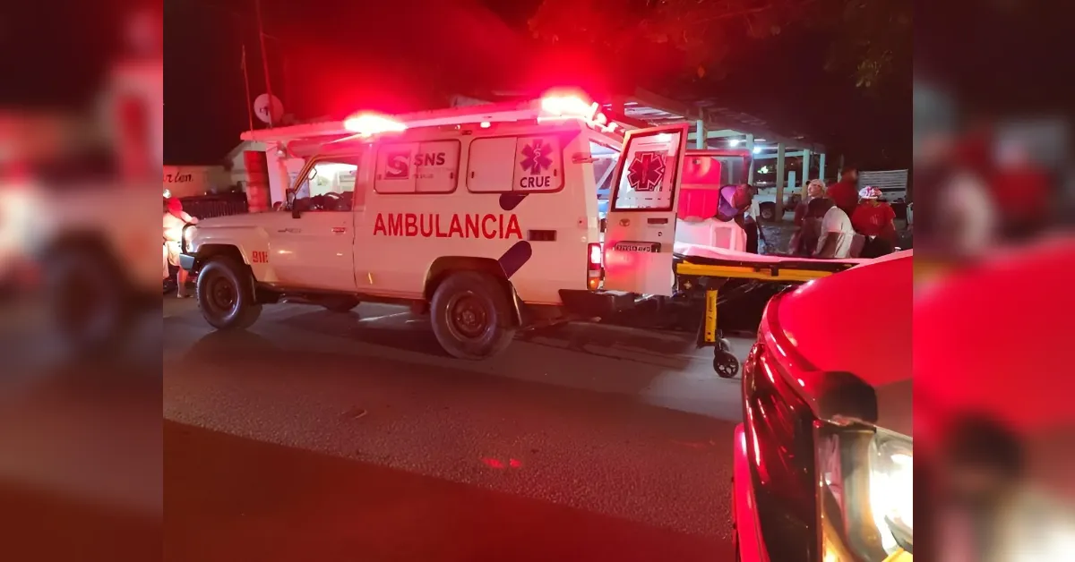 Ambulance with red flashing lights at night, rear doors open and a stretcher nearby, waiting on a street with people around.