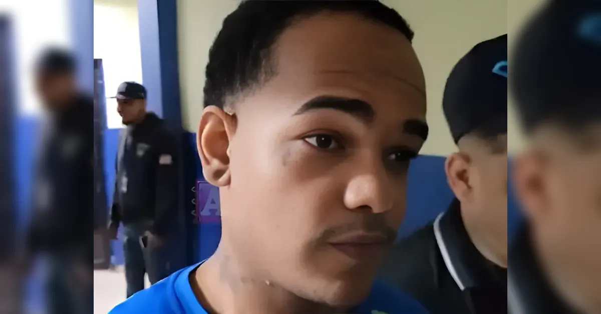 Close-up of a man in a blue shirt, looking to the left, with blurred people in a hallway behind him.