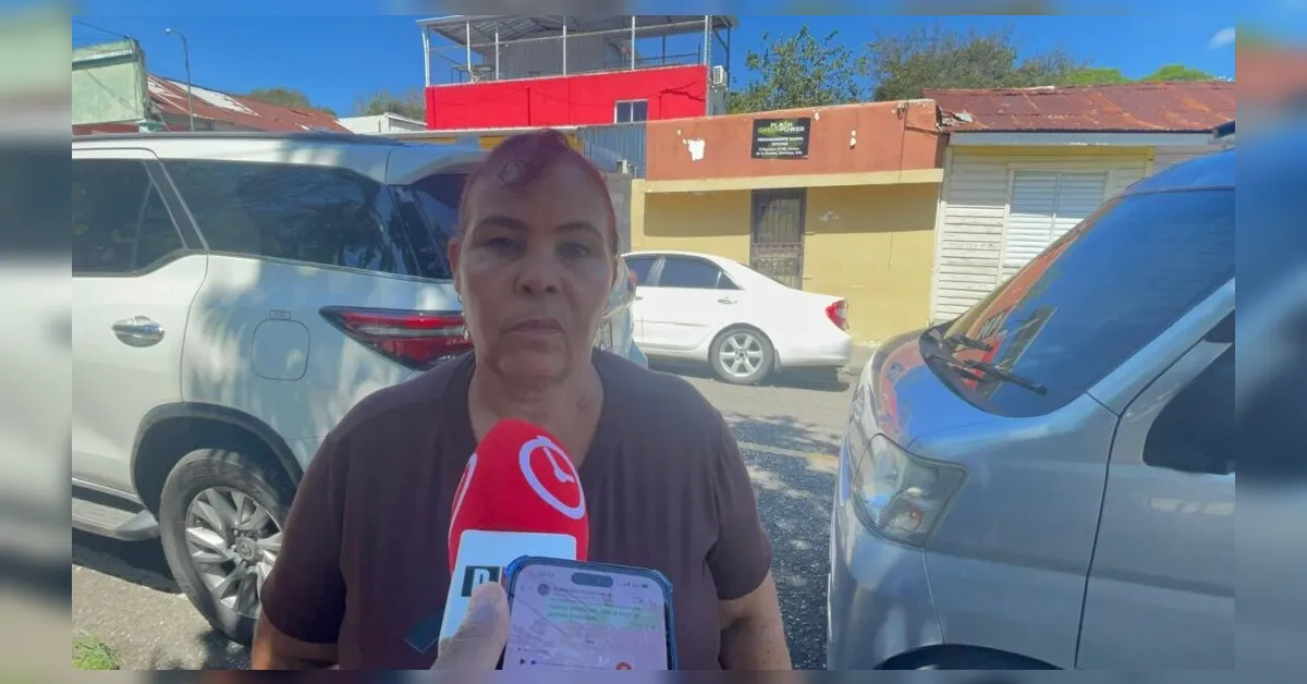 Woman being interviewed on a sunny street, microphone held near her face with reporters nearby, cars parked in the background