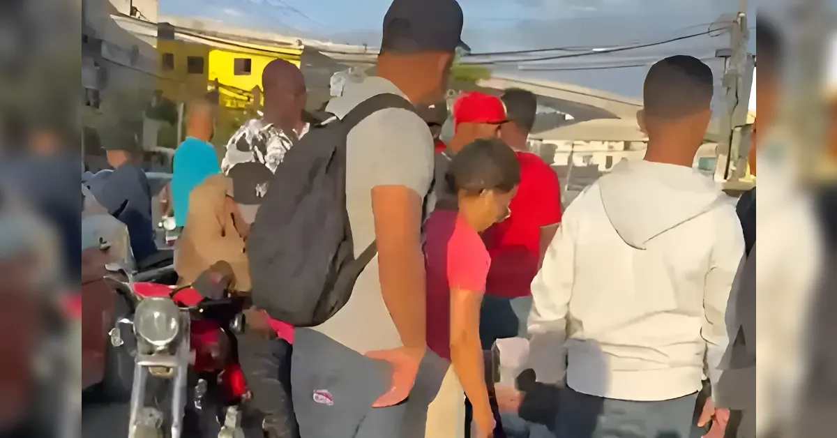 Group of people standing and talking on a sunlit street; a man with a backpack sits beside a motorcycle in the foreground