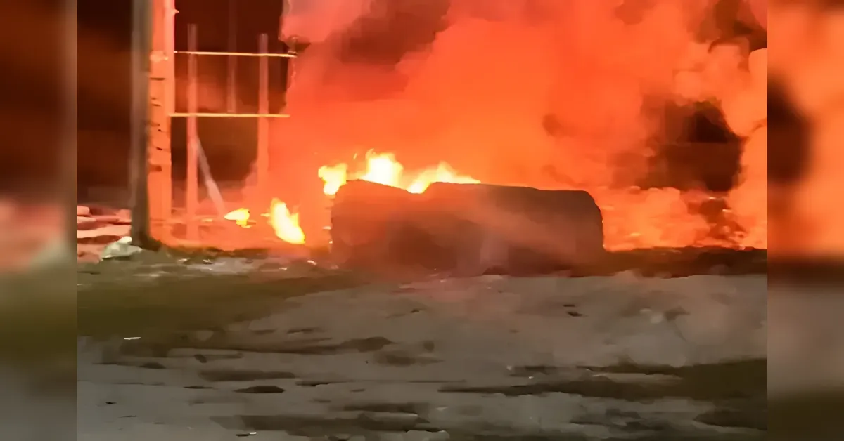 A large outdoor fire with bright orange flames engulfing a dark, cylindrical object at night.