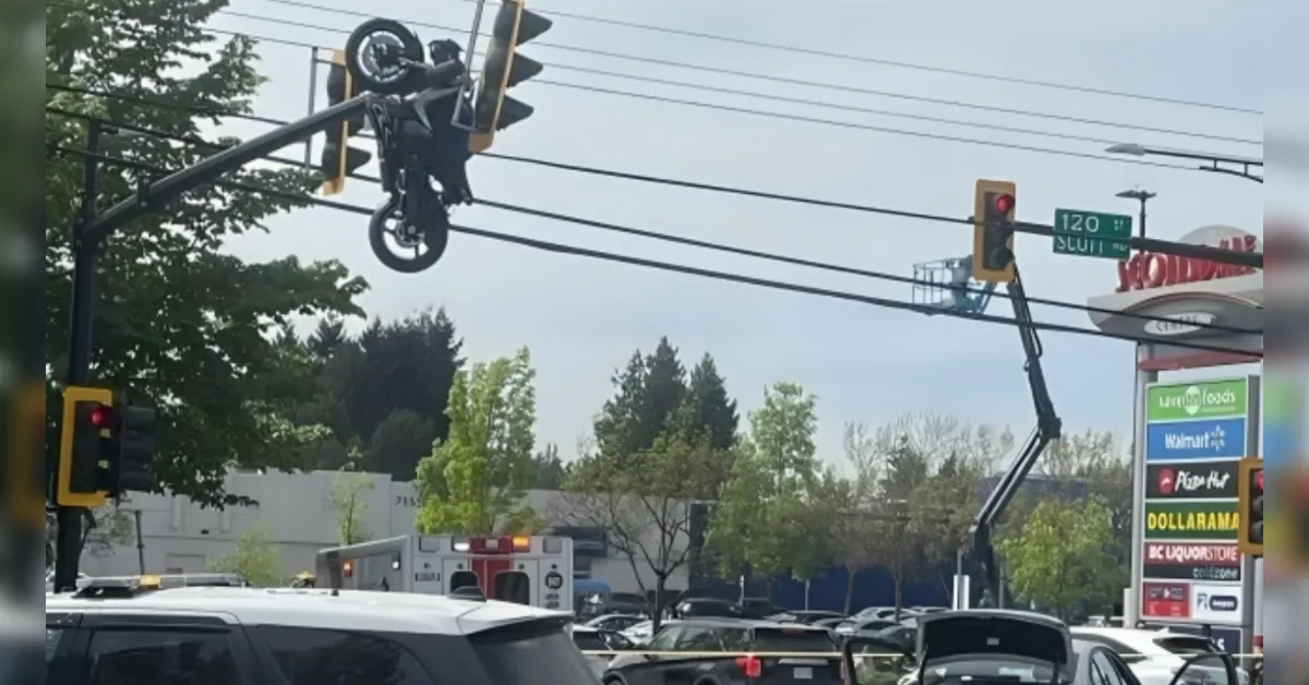 Motorcycle tangled in power lines above a busy intersection, with a crane arm lifting it while cars and an ambulance are visible below.