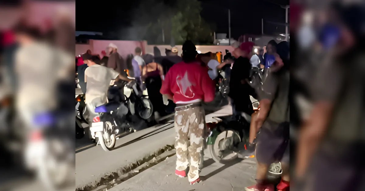 Nighttime street scene with a crowd gathered around parked motorbikes and scooters on a dimly lit sidewalk, silhouettes and clothing visible in motion.