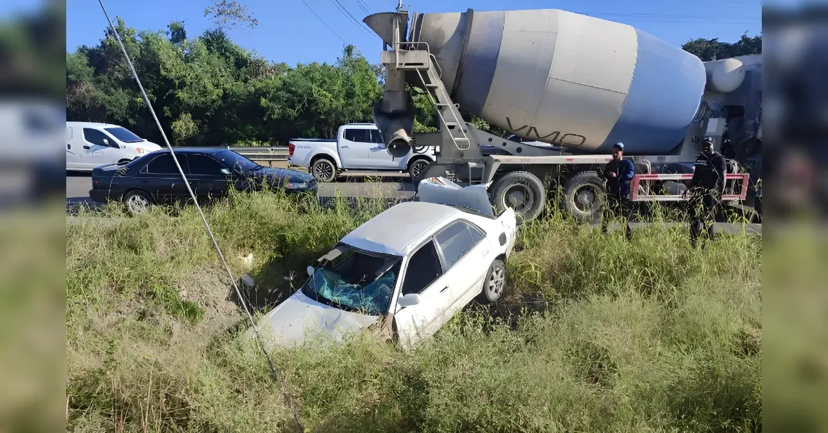 White sedan in a grassy ditch beside a large cement mixer truck on a highway, with bystanders and workers nearby on a sunny day.