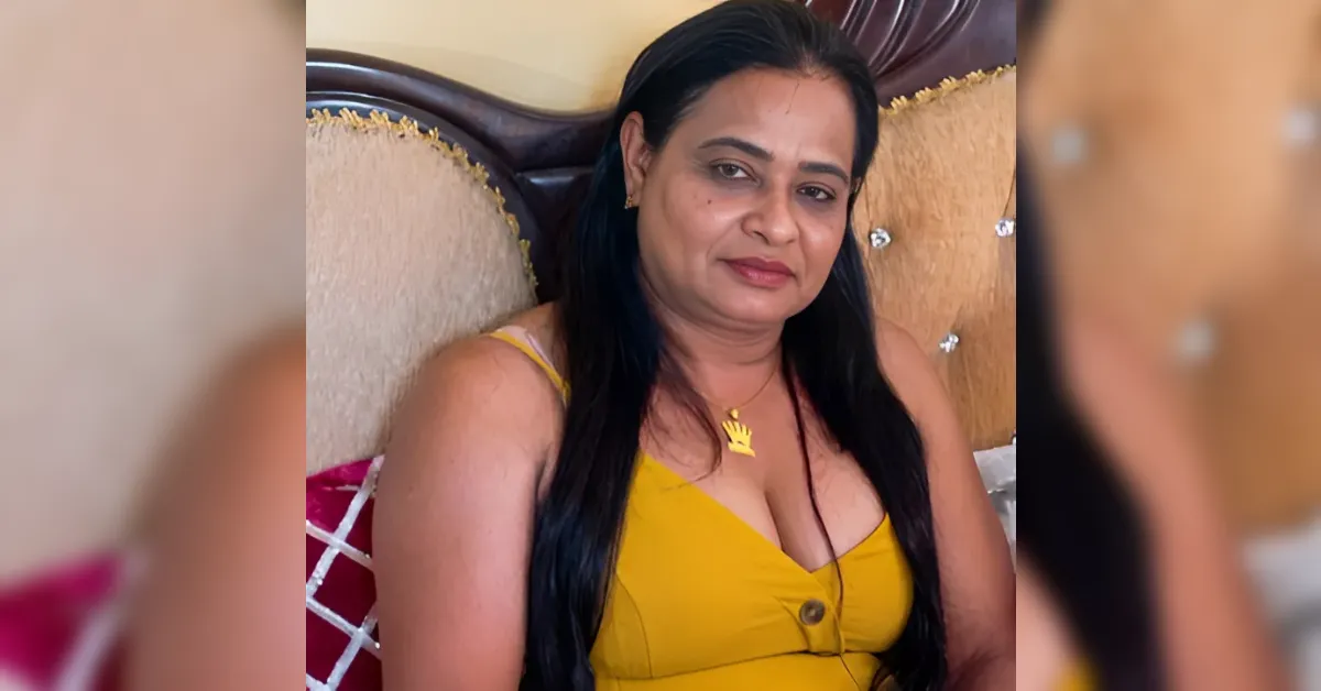 Woman with long black hair in a mustard yellow sleeveless top sits on a tufted couch, looking at the camera with a slight smile.