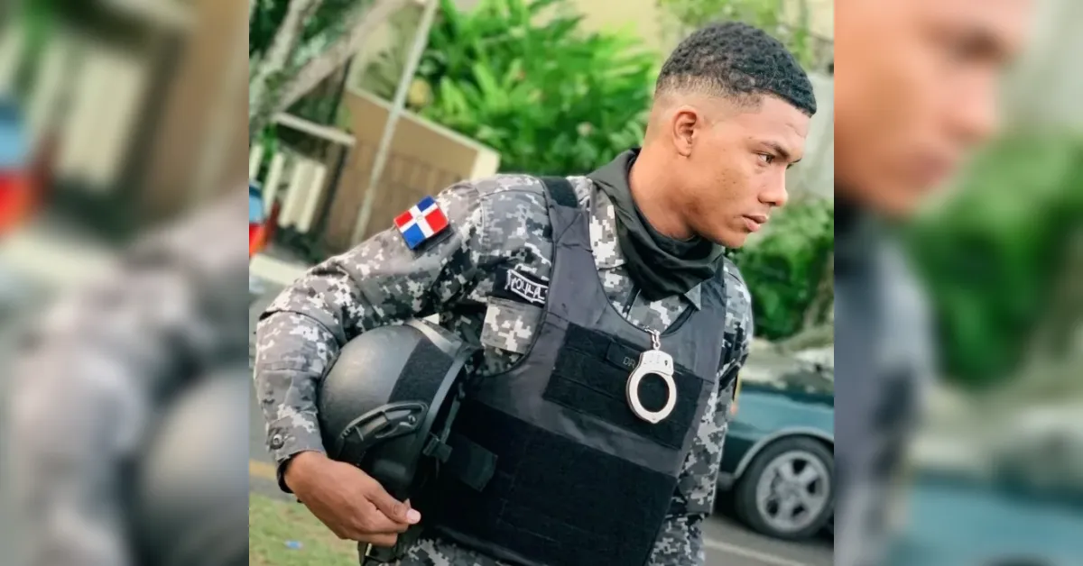 Young man in camouflage tactical gear, holding a helmet, wearing a bulletproof vest with a metal handcuff and a Dominican flag patch on the sleeve.