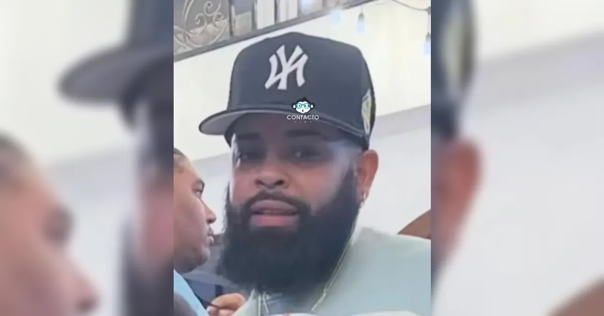 Close-up of a bearded man wearing a black baseball cap with a white NY logo, speaking in a crowded indoor setting, smoke visible near his face.